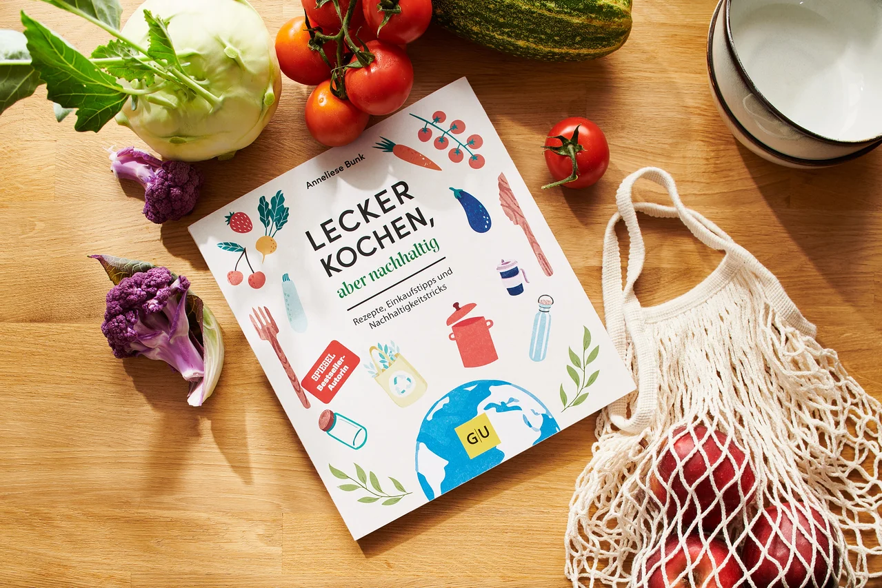 Kochbuch »Lecker kochen, aber nachhaltig« liegt auf Holztisch mit Tomaten, Blumenkohl, Kohlrabi, Äpfeln in Stoffbeutel und Schüsseln.
