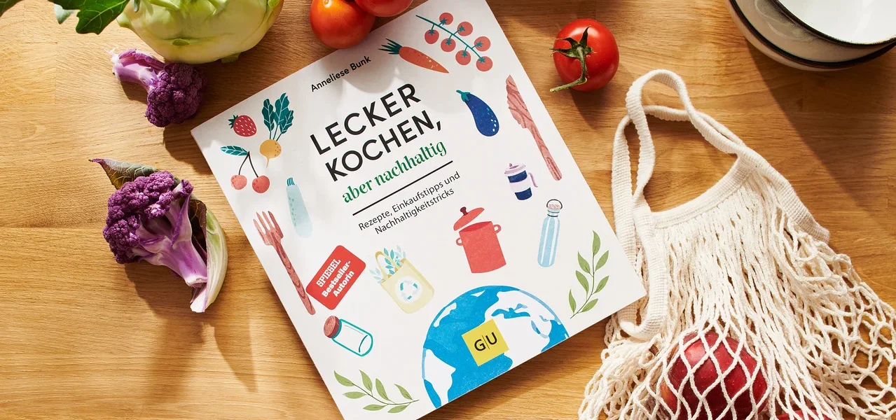 Kochbuch »Lecker kochen, aber nachhaltig« liegt auf Holztisch mit Tomaten, Blumenkohl, Kohlrabi, Äpfeln in Stoffbeutel und Schüsseln.