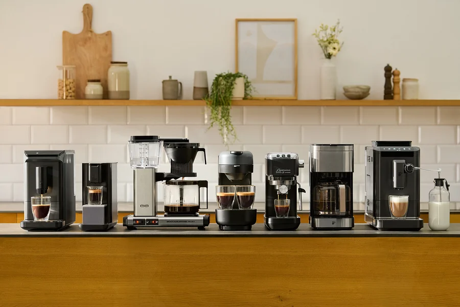 Verschiedene Kaffeemaschinen stehen auf einer Küchentheke vor einer weißen Fliesenkulisse.