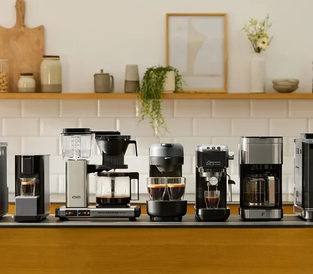 Verschiedene Kaffeemaschinen stehen auf einer Küchentheke vor einer weißen Fliesenkulisse.