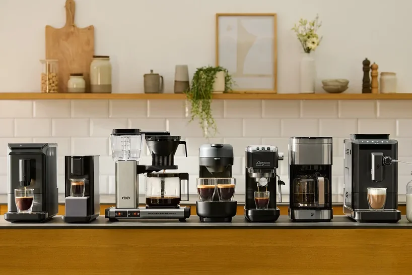 Verschiedene Kaffeemaschinen stehen auf einer Küchentheke vor einer weißen Fliesenkulisse.