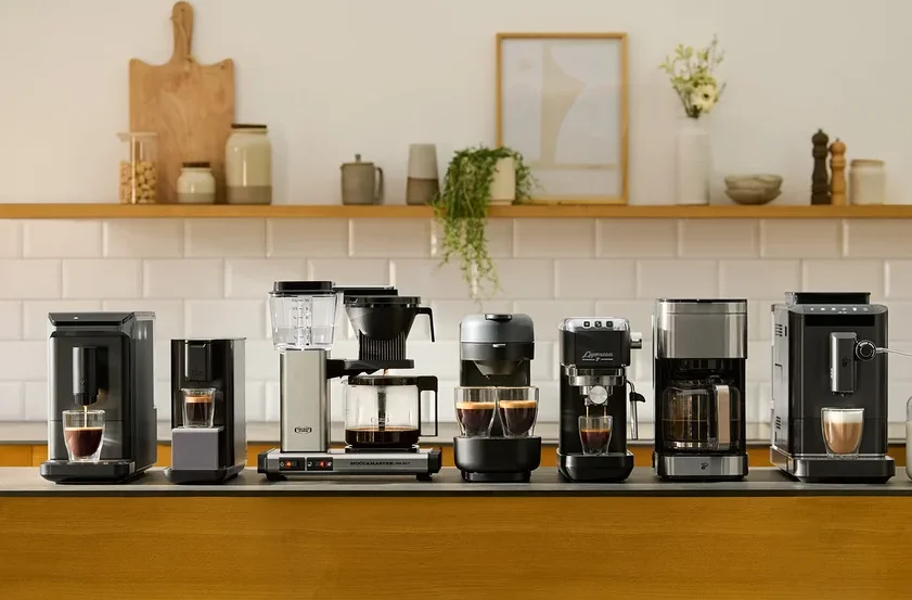 Verschiedene Kaffeemaschinen stehen auf einer Küchentheke vor einer weißen Fliesenkulisse.