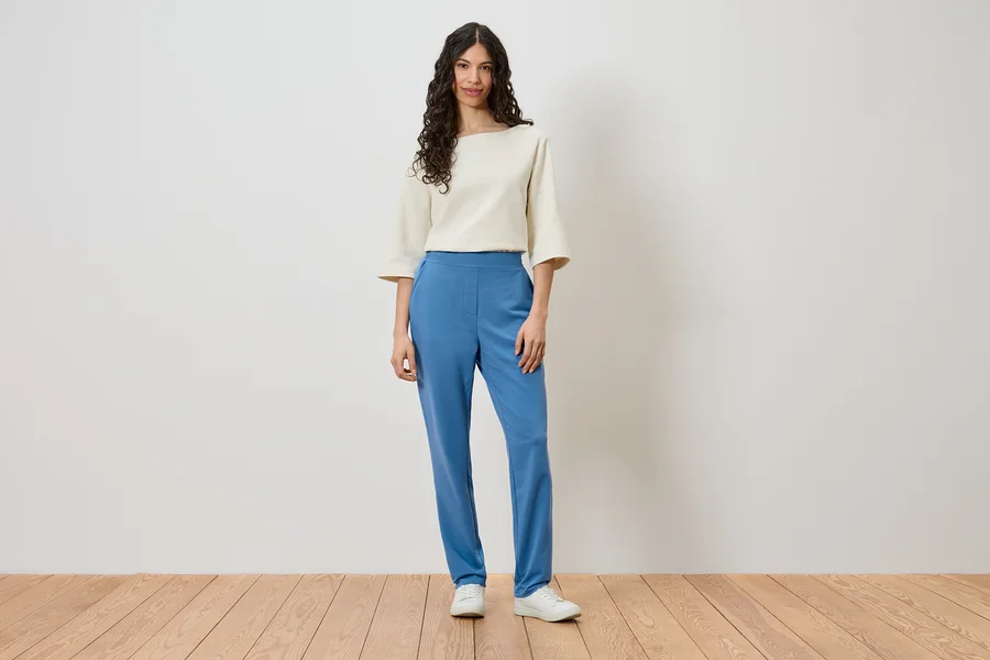 Eine Frau steht vor einer Wand und trägt ein cremefarbenes Oberteil, blaue Hosen und weiße Schuhe.