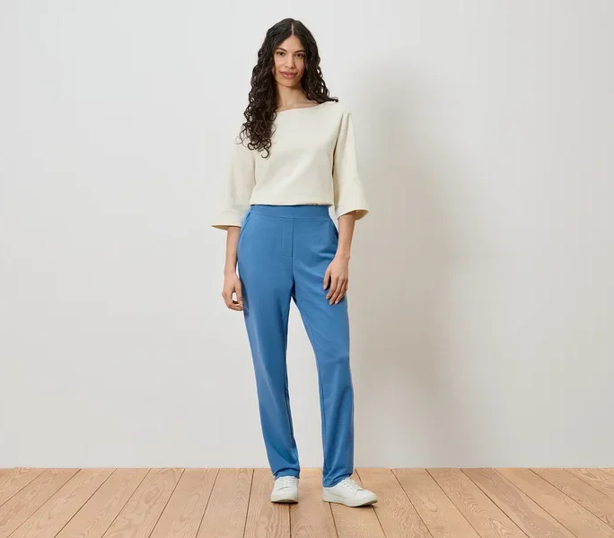 Eine Frau steht vor einer Wand und trägt ein cremefarbenes Oberteil, blaue Hosen und weiße Schuhe.
