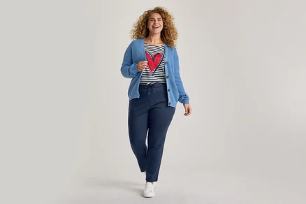 Eine lächelnde Frau mit lockigem Haar trägt einen blauen Strickcardigan, ein gestreiftes T-Shirt mit rotem Herz, dunkelblaue Jogginghose und weiße Sneaker, während sie nach vorne geht.