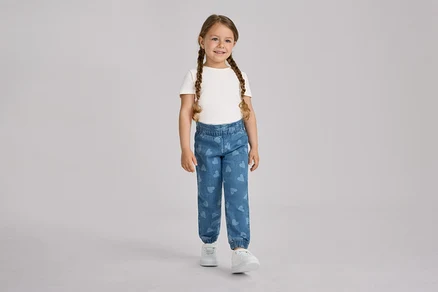 Ein junges Mädchen mit zwei Zöpfen steht lächelnd da und trägt ein weißes T-Shirt sowie blaue Kinder-Pull-on-Pants – Fit »Ella« mit Herzmustern und weiße Turnschuhe.