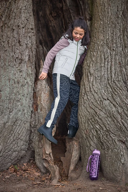 Mädchen klettert in einen hohlen Baumstamm, trägt eine helle Weste, gemusterte Hose und dunkelblaue Kinder-Regen-Chelseaboots. Eine violette Trinkflasche liegt am Boden.