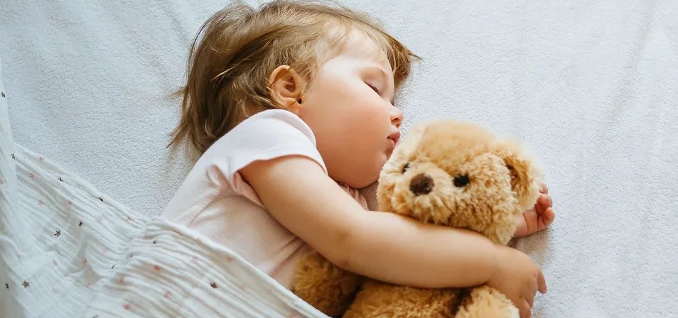 Ein schlafendes Kind umarmt einen Teddybären unter einer Decke auf einem weißen Bettlaken.