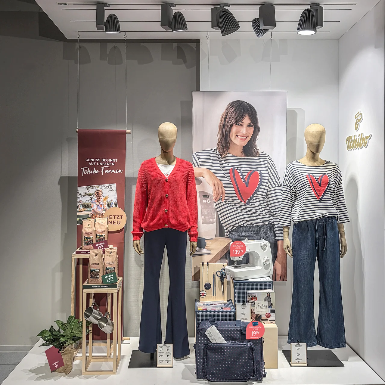 Schaufensterauslage mit zwei Schaufensterpuppen. Die linke trägt einen roten Cardigan und dunkelblaue Schlaghosen, die rechte ein gestreiftes Oberteil mit rotem Herz und blaue Jeans. Dahinter ein Poster einer lächelnden Frau. Links sind Kaffeeprodukte ausgestellt, rechts eine Nähmaschine und Nähzubehör.