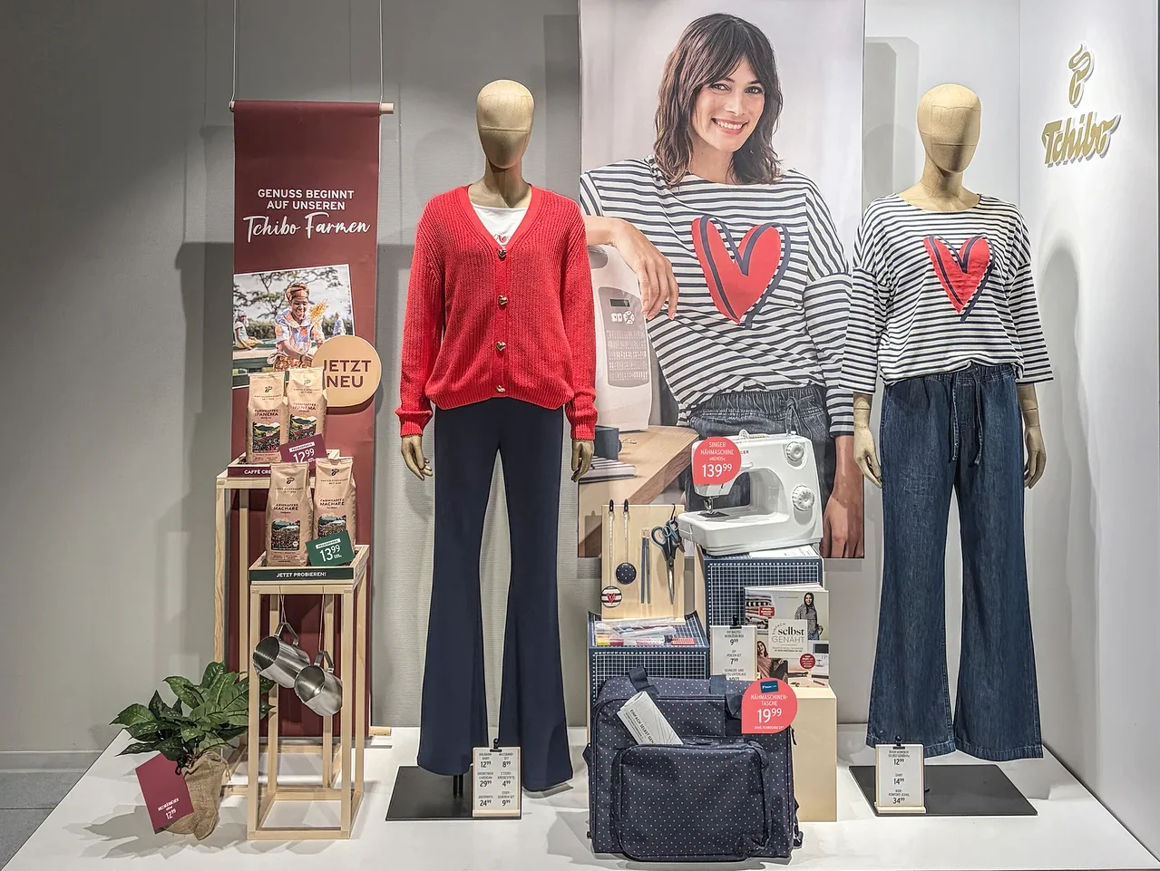 Schaufensterauslage mit zwei Schaufensterpuppen. Die linke trägt einen roten Cardigan und dunkelblaue Schlaghosen, die rechte ein gestreiftes Oberteil mit rotem Herz und blaue Jeans. Dahinter ein Poster einer lächelnden Frau. Links sind Kaffeeprodukte ausgestellt, rechts eine Nähmaschine und Nähzubehör.