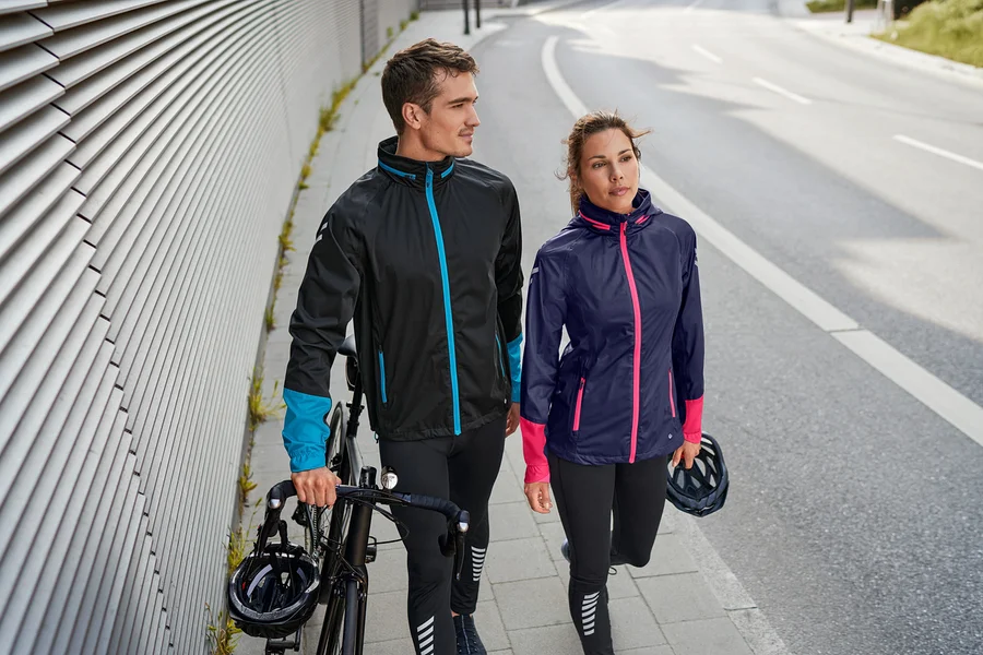 Ein Mann und eine Frau gehen auf dem Bürgersteig. Der Mann hält ein Fahrrad und trägt eine Fahrrad-Regenjacke und eine Fahrradhose. Die Frau trägt eine Fahrrad-Regenjacke und eine Fahrradhose.