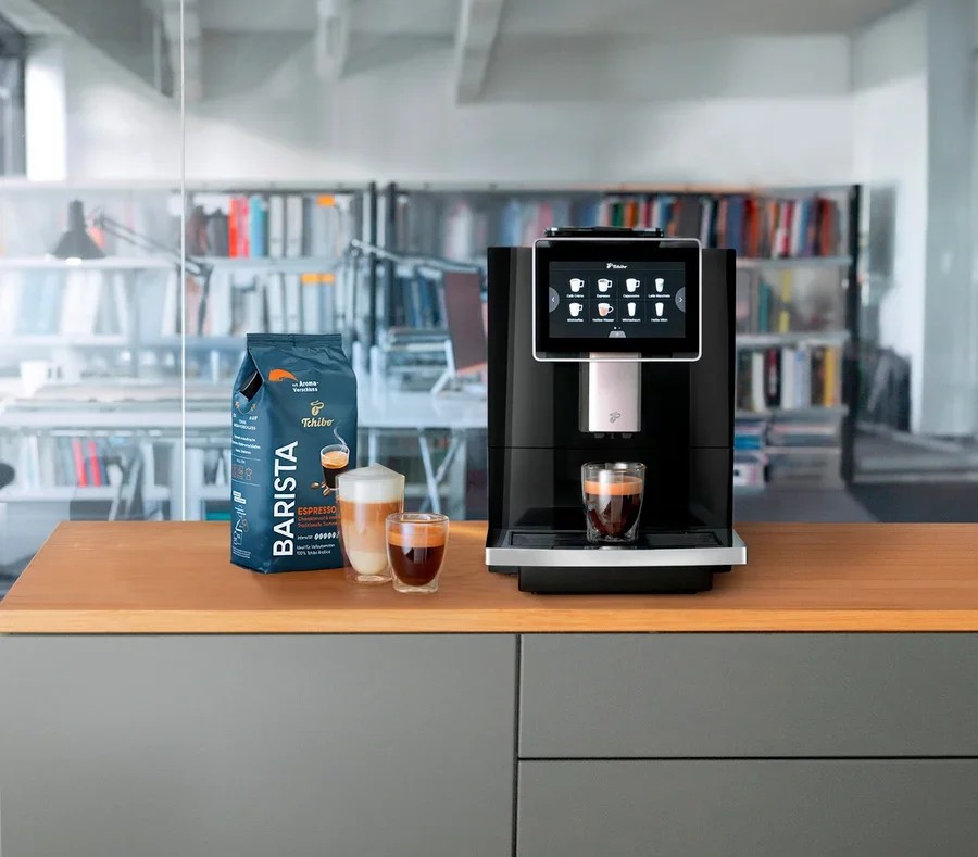 Ein Kaffeevollautomat steht neben einer Packung Barista-Kaffee und zwei Gläsern Kaffee.