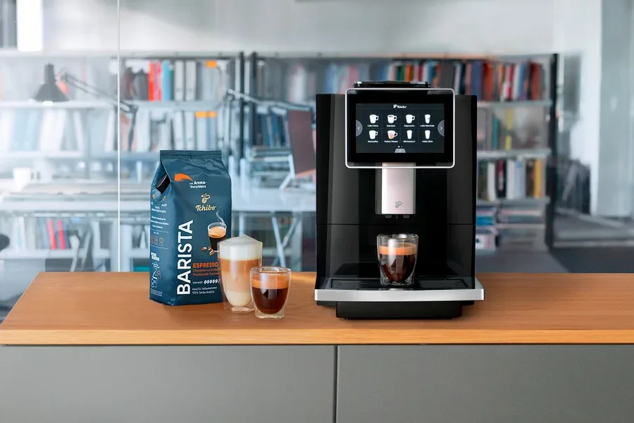 Ein Kaffeevollautomat steht neben einer Packung Barista-Kaffee und zwei Gläsern Kaffee.