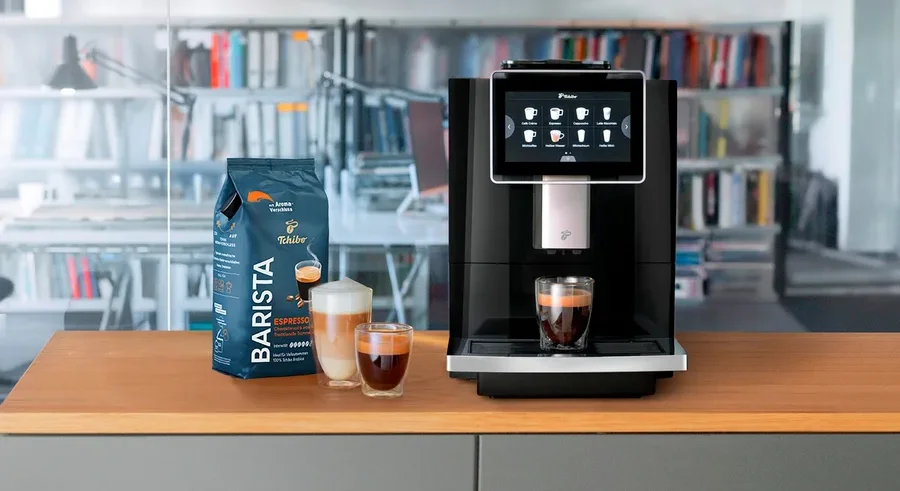 Ein Kaffeevollautomat steht neben einer Packung Barista-Kaffee und zwei Gläsern Kaffee.