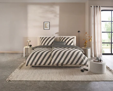 Dunkelgrau-beige gestreifte SCHÖNER WOHNEN-Kollektion Renforcé-Bettwäsche auf einem Bett in einem modernen Schlafzimmer mit hellem Teppich und Fenster.