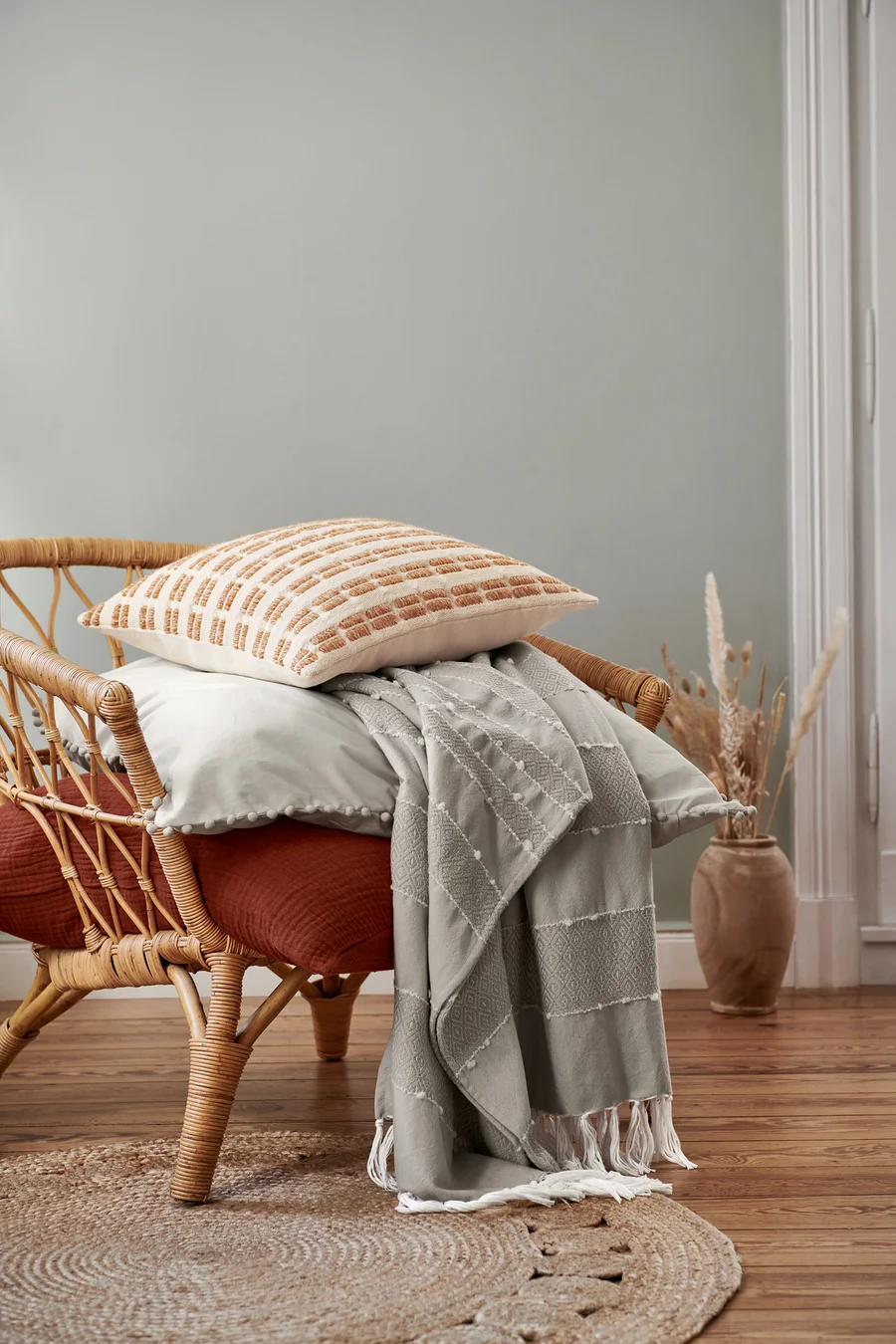 Ein beiger Dekokissen und eine Musselin-Bettwäsche in Normalgröße liegen auf einem Rattansessel. Darüber ist eine Wohndecke drapiert.