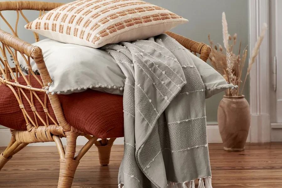 Ein beiger Dekokissen und eine Musselin-Bettwäsche in Normalgröße liegen auf einem Rattansessel. Darüber ist eine Wohndecke drapiert.