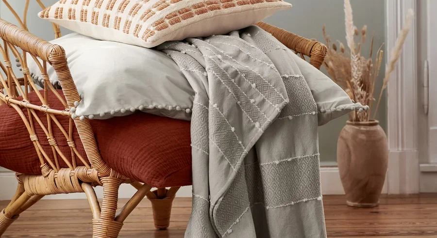 Ein beiger Dekokissen und eine Musselin-Bettwäsche in Normalgröße liegen auf einem Rattansessel. Darüber ist eine Wohndecke drapiert.