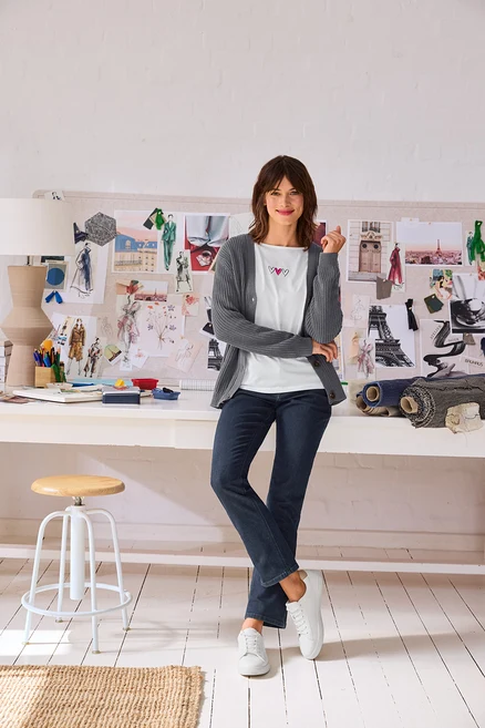 Lächelnde Frau in grauem Cardigan, weißem T-Shirt mit Herzen, dunklen Jeans und weißen Leder-Sneakern, trägt Gold-Doppelcreolen und steht vor einem Schreibtisch mit Moodboard.