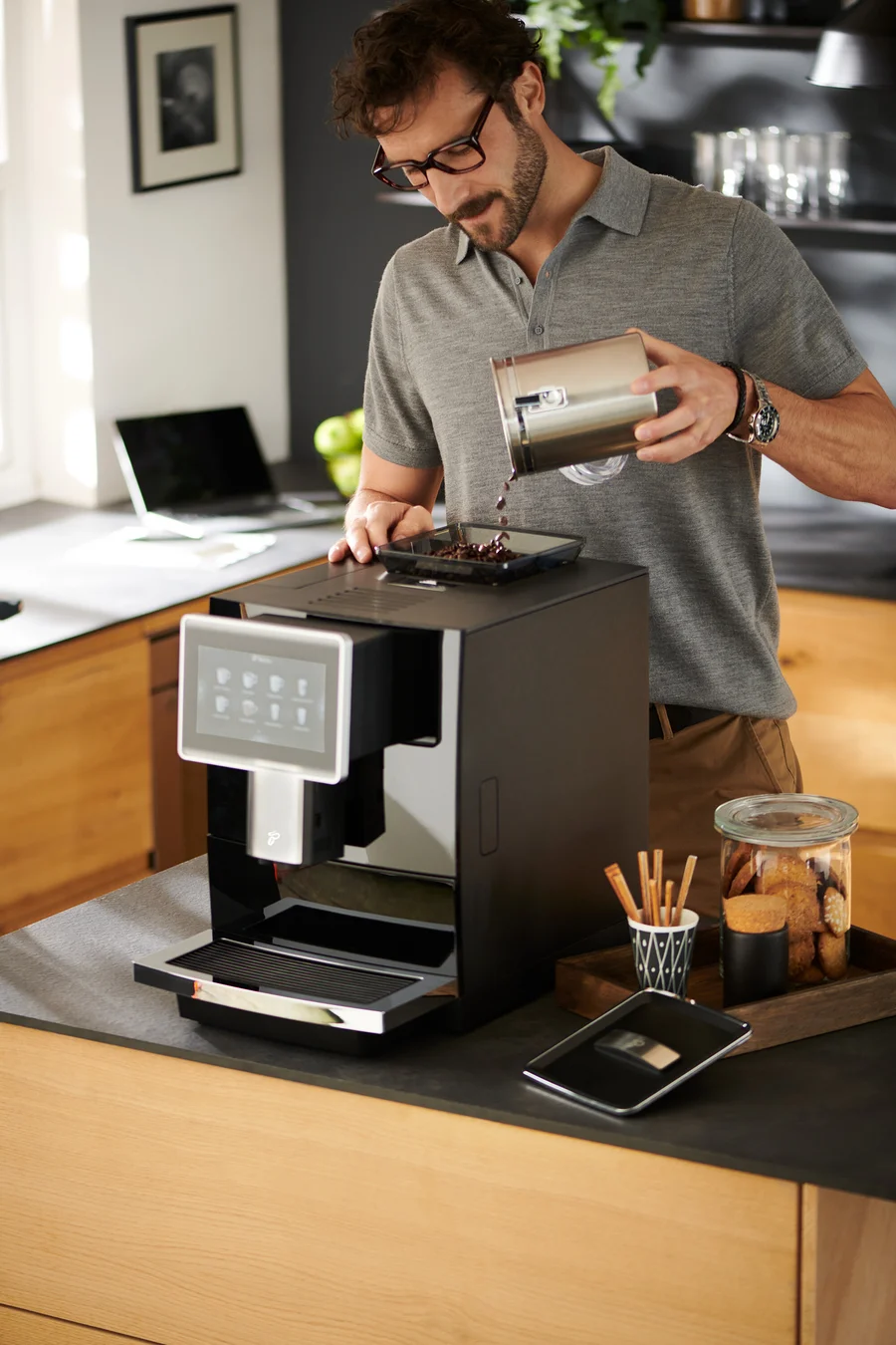 Mann mit Brille gießt Kaffeebohnen in den »Tchibo Office« Kaffeevollautomat, schwarz.