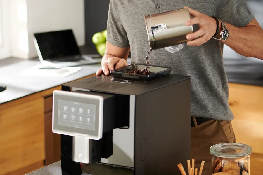 Mann mit Brille gießt Kaffeebohnen in den »Tchibo Office« Kaffeevollautomat, schwarz.