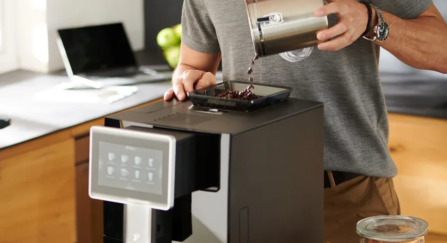 Mann mit Brille gießt Kaffeebohnen in den »Tchibo Office« Kaffeevollautomat, schwarz.