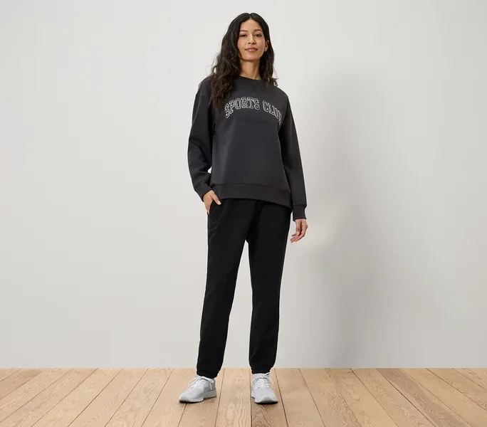 Frau steht mit Händen in den Taschen ihrer schwarzen Jogginghose. Sie trägt ein graues Sweatshirt mit der Aufschrift Sports Club und weiße Turnschuhe.