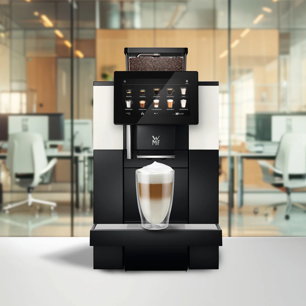 Ein Kaffeevollautomat mit Touchscreen, der Getränkeoptionen anzeigt, und einem Bohnenbehälter voller Kaffeebohnen. Ein Glas geschichteter Latte Macchiato steht auf der Abtropfschale, im Hintergrund ein verschwommenes Büro.