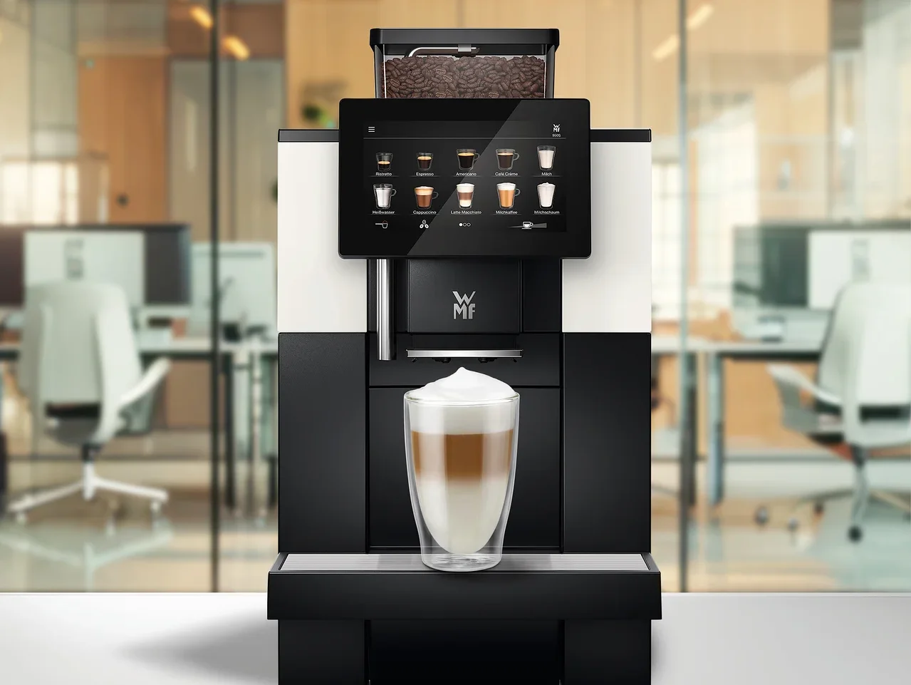 Ein Kaffeevollautomat mit Touchscreen, der Getränkeoptionen anzeigt, und einem Bohnenbehälter voller Kaffeebohnen. Ein Glas geschichteter Latte Macchiato steht auf der Abtropfschale, im Hintergrund ein verschwommenes Büro.