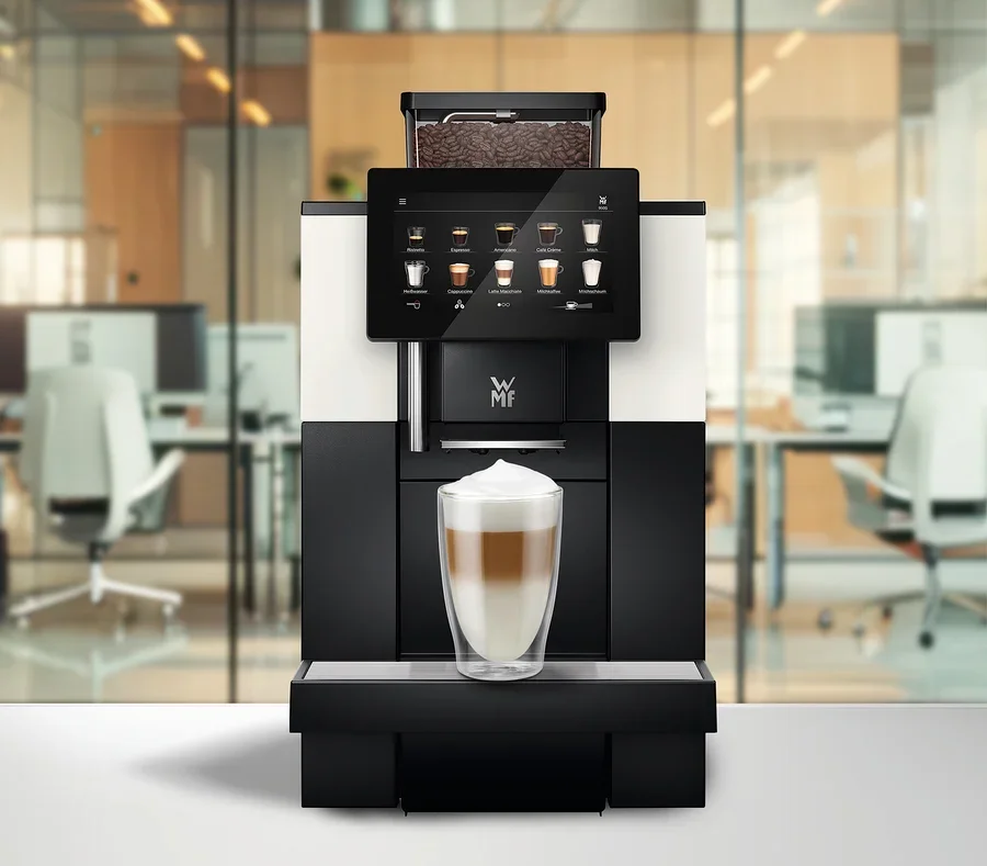 Ein Kaffeevollautomat mit Touchscreen, der Getränkeoptionen anzeigt, und einem Bohnenbehälter voller Kaffeebohnen. Ein Glas geschichteter Latte Macchiato steht auf der Abtropfschale, im Hintergrund ein verschwommenes Büro.