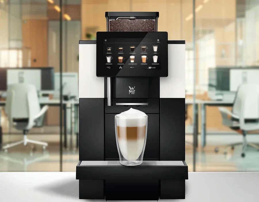 Ein Kaffeevollautomat mit Touchscreen, der Getränkeoptionen anzeigt, und einem Bohnenbehälter voller Kaffeebohnen. Ein Glas geschichteter Latte Macchiato steht auf der Abtropfschale, im Hintergrund ein verschwommenes Büro.