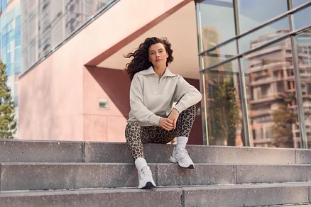 Frau mit lockigem Haar sitzt auf Betonstufen, trägt ein beiges Oberteil mit halbem Reißverschluss, Leggings mit Leopardenmuster und weiße Turnschuhe, im Hintergrund moderne Gebäude.