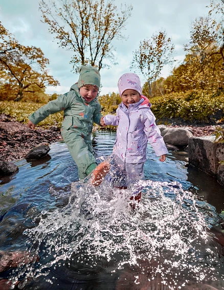 Zwei kleine Kinder in grünen und lila wasserdichten Anzügen planschen im flachen Bach, halten Händchen, mit Herbstbäumen im Hintergrund.