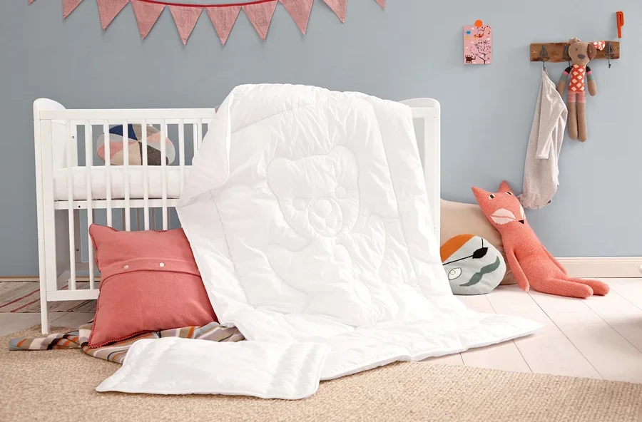 Weiße Steppdecke mit Kissen und rosa Dekokissen neben einem weißen Kinderbett.