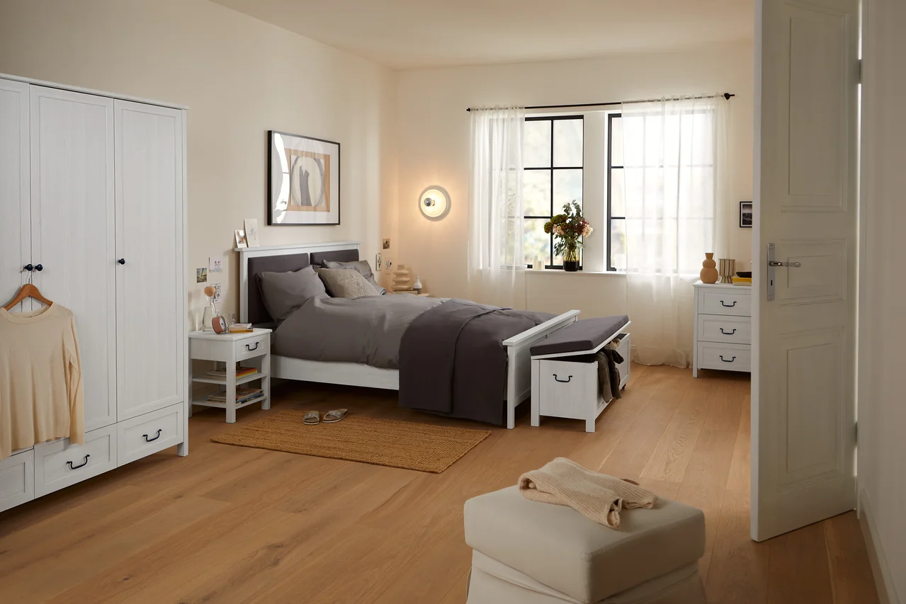Schlafzimmer mit Bett mit gepolstertem Kopfteil, Kommode mit 3 Schubladen, Kleiderschrank, 3-Türer, Sitzbank mit Truhenfunktion und Nachttisch mit Schublade.