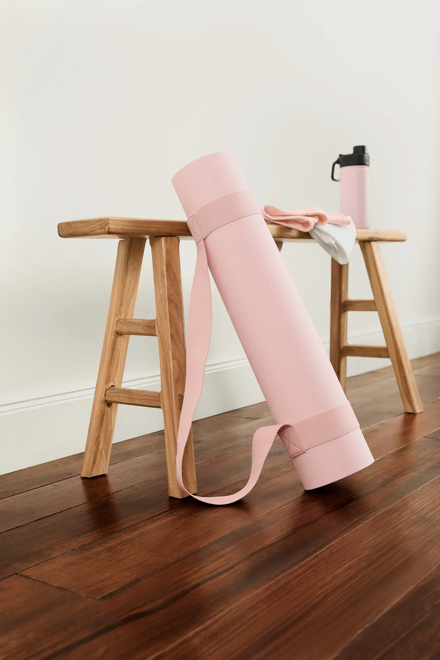 Eine zusammengerollte, rosa Yoga-und-Fitness-Matte lehnt an einem Holzhocker. Auf dem Hocker liegen ein gefaltetes Handtuch und eine rosa Wasserflasche.