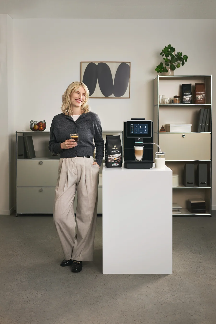 Frau hält ein Glas Kaffee neben einer Tchibo Espresso Maschine und einer Packung Espresso.