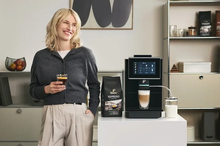 Frau hält ein Glas Kaffee neben einer Tchibo Espresso Maschine und einer Packung Espresso.