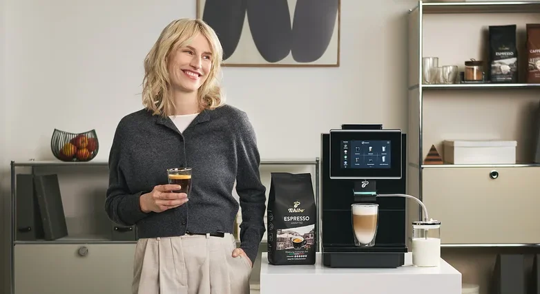 Frau hält ein Glas Kaffee neben einer Tchibo Espresso Maschine und einer Packung Espresso.