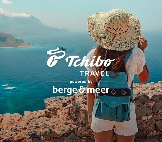 Frau mit Hut und blauem Rucksack steht auf Felsen mit Blick aufs Meer. Logos von Tchibo Travel und Berge&Meer.