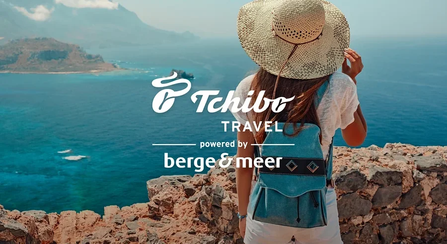 Frau mit Hut und blauem Rucksack steht auf Felsen mit Blick aufs Meer. Logos von Tchibo Travel und Berge&Meer.