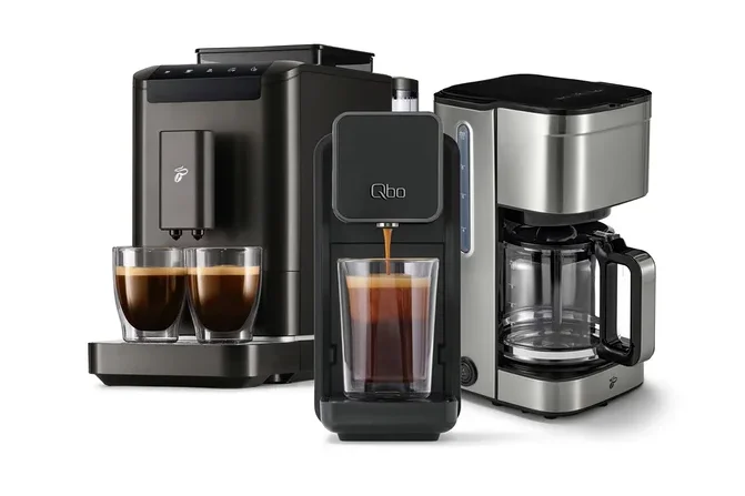 Drei Kaffeemaschinen: links eine dunkelgraue Espressomaschine mit zwei gefüllten Gläsern, mittig eine schwarze Qbo-Kapselmaschine, die Kaffee in ein Glas füllt, und rechts eine silberne Filterkaffeemaschine.