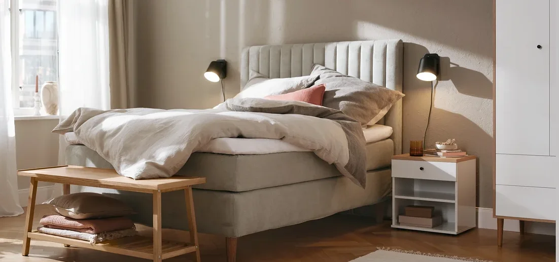 Beiges Boxspringbett (ca. 180 x 200 cm), Kleiderschrank, Nachttisch namens »Bekeson«, schwarze Wandleuchte aus Metall und Sitzbank mit Ablage.