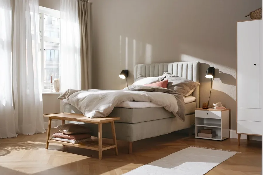 Beiges Boxspringbett (ca. 180 x 200 cm), Kleiderschrank, Nachttisch namens »Bekeson«, schwarze Wandleuchte aus Metall und Sitzbank mit Ablage.