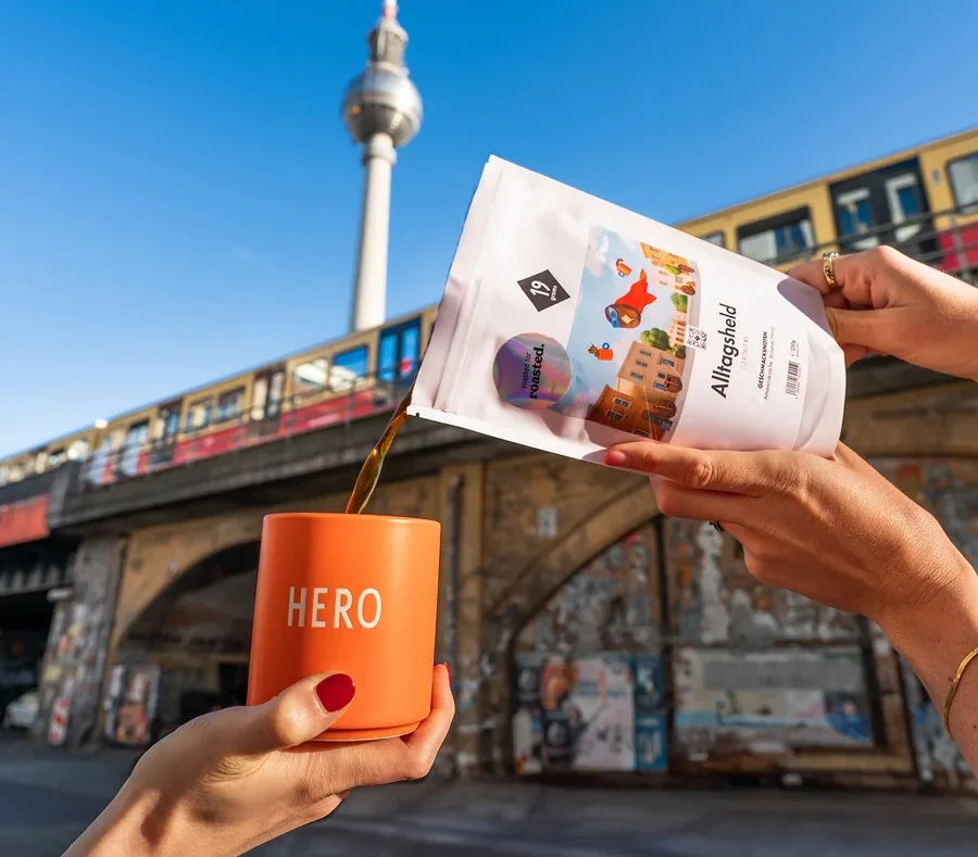 Eine Person gießt Kaffee aus einer weißen Packung in eine orangefarbene Tasse mit der Aufschrift HERO, im Hintergrund der Berliner Fernsehturm.