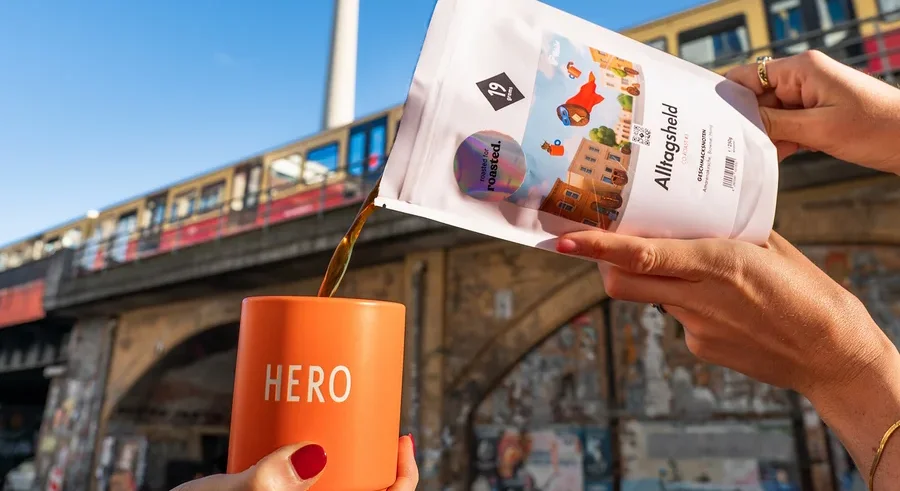 Eine Person gießt Kaffee aus einer weißen Packung in eine orangefarbene Tasse mit der Aufschrift HERO, im Hintergrund der Berliner Fernsehturm.