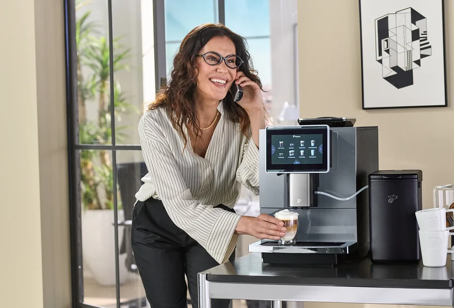Frau hält ein Glas Kaffee an einer Kaffeemaschine und telefoniert.