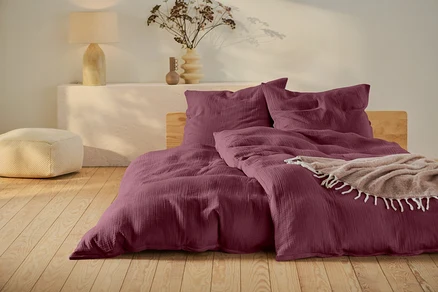 Weinrote Bettwäsche auf einem Bett mit Holzrahmen, ein beiger Pouf und eine Lampe auf einem weißen Tisch.