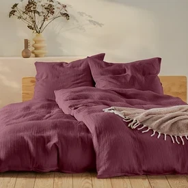 Weinrote Bettwäsche auf einem Bett mit Holzrahmen, ein beiger Pouf und eine Lampe auf einem weißen Tisch.