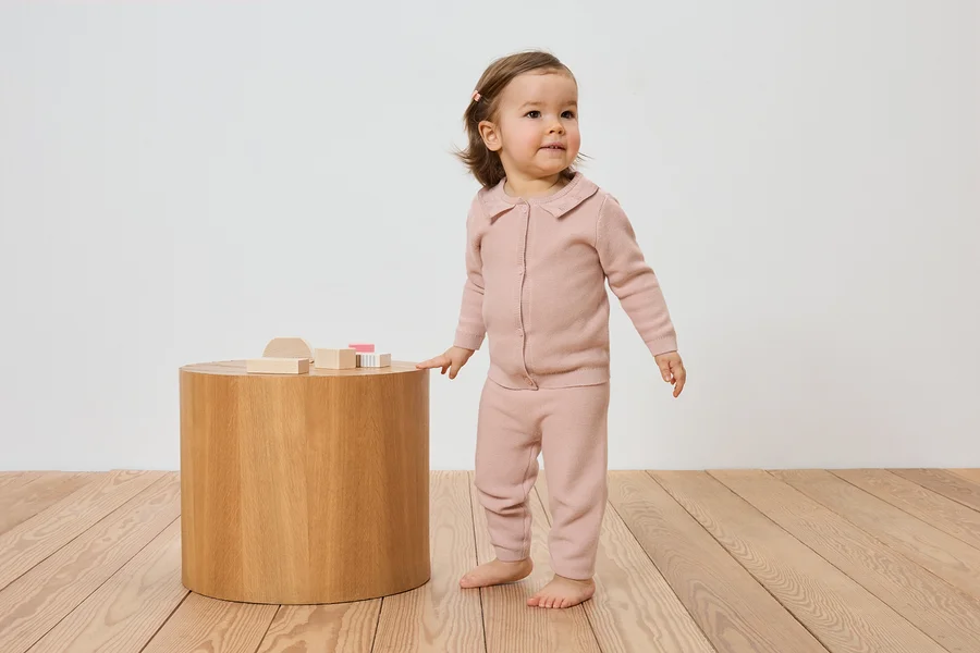 Kleinkind in rosa Strickjacke und Hose steht neben einem Holztisch mit Holzspielzeug auf einem Holzboden.